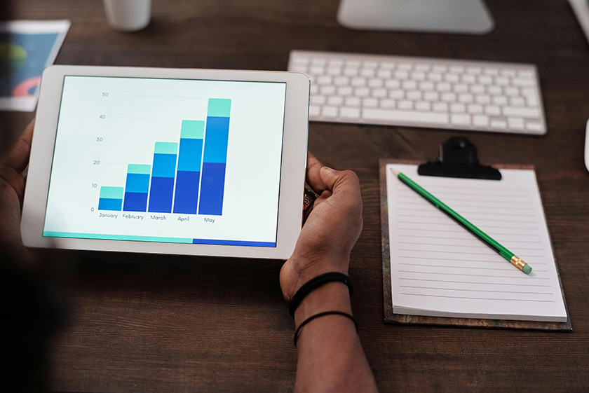 Persona sosteniendo una tablet que muestra un gráfico de barras con el crecimiento de datos de enero a mayo, junto a un portapapeles con un bloc de notas y un lápiz verde sobre un escritorio.