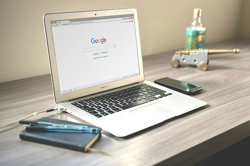 MacBook Air abierto mostrando la página de inicio de Google sobre un escritorio de madera, con un smartphone y otros elementos decorativos cerca.